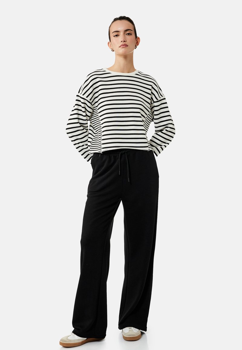 Femme debout les mains dans les poches portant une chemise à manches longues blanche et noire à rayures, un pantalon large noir et des baskets blanches.