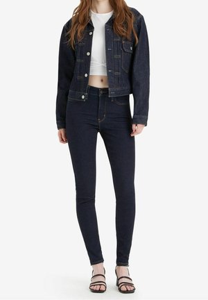 Vrouw met een donker denimjack en spijkerbroek, wit cropped topje en zwarte sandalen met bandjes tegen een effen achtergrond.