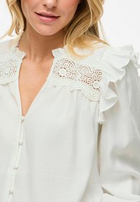 Bílá blůza s krajkovými detaily na výstřihu, volánkovými akcenty na ramenou a malými knoflíkovými zapínáními vpředu. Měkčená, lehká tkanina.