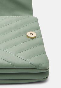 Cartera de cuero verde con acolchado y textura suave, con un detalle de hardware dorado redondo y costuras visibles en un tono contrastante.