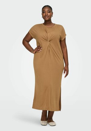 Tumejume nahaga naine, kes kannab beežikat lühikeste varrukatega, pahkluuni ulatuvat kleiti, millel on ees sõlme detail, seisab ühe käega puusal ja seljas beežid platvormkingad.