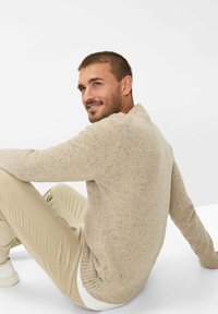 Beige Strickpullover mit Rundhalsausschnitt, der in verschiedenen Farben gesprenkelt ist. Gerippte Bündchen und Saum, kombiniert mit hellbraunen Hosen.