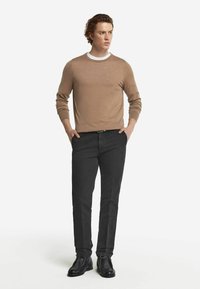 Beige, långärmad tröja med vit halsringning, kombinerad med svarta, raka byxor och svarta snörskor, stående mot en neutral bakgrund.