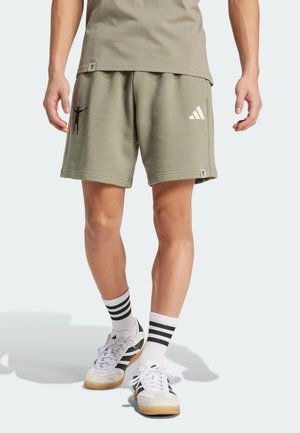 Personne portant un short vert olive avec des logos, des chaussettes blanches à rayures noires et des chaussures de sport blanches, debout devant un fond uni.