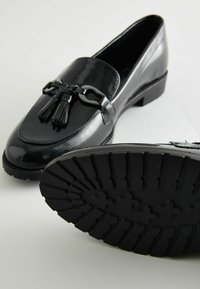 Schwarze lacklederne Slipper verfügen über eine Ketten- und Quastenakzentuierung, einen niedrigen Absatz und eine strukturierte Gummisohle mit einem gemusterten Profil.