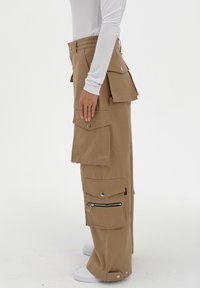 Baggy beige cargobroeken met meerdere grote zakken, zilverkleurige hardware en een ritszak aan de zijkant. Gemaakt van duurzaam materiaal.