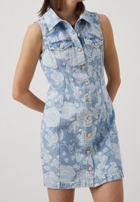 Denim dress in light blue with a floral pattern. Features sleeveless design, button front, and two chest pockets. Silhouette is fitted.
