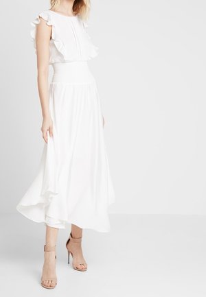 Une femme se tient en souriant dans une robe blanche fluide avec des manches froncées et une taille cintrée, accompagnée de sandales beiges à lanières.