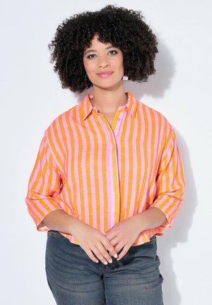 Vrouw met krullend haar die een roze en oranje shirt met verticale strepen en een blauwe spijkerbroek draagt, staand met de handen gevouwen.