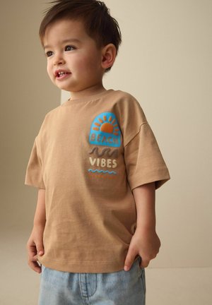 Jonge jongen die een beige T-shirt draagt met een zon, golven en de tekst "Beach Vibes", gecombineerd met lichtblauwe denim shorts, staand tegen een effen achtergrond.