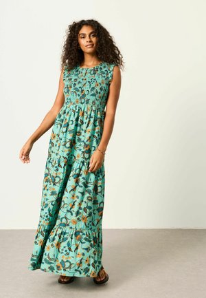 Kvinde med krøllet hår iført en ærmeløs teal blomster maxi kjole og sandaler mod en ensfarvet lys baggrund.