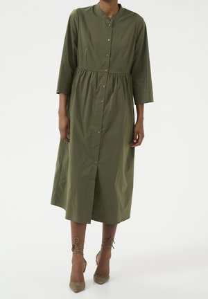 Kobieta ubrana w oliwkową sukienkę midi na guziki z rękawami 3/4 oraz beżowe szpilki z wiązaniami wokół kostek.