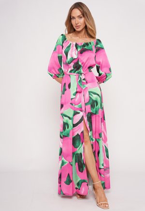 Woman wearing a colorful pink, green, and black patterned long dress with puffed sleeves and a high slit, standing against a plain background.