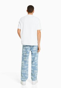 Vit kortärmad skjorta i kombination med ljusblå jeans med ett upprepande mönster av vit graffiti-liknande text. Sneakers fulländar looken.