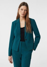 Woman with long brown hair wearing teal blazer and matching pants over a black top, standing with one hand in pocket on white background.