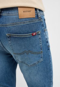 Niebieskie dżinsy z denimu z skórzaną metką z napisem "MUSTANG" na pasku, czerwoną metką na tylnej kieszeni i żółtymi przeszyciami.