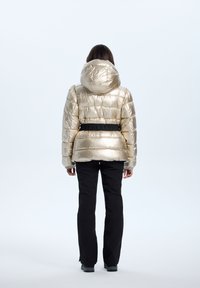 Guldmetalisk pufferjakke med hætte, elastiske manchetter og et sort bælte i taljen; båret med sorte bukser; glat, skinnende tekstur.