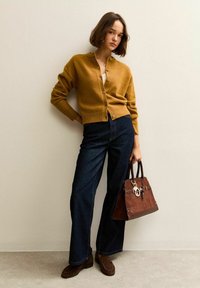 Musztardowy kardigan z guzikami, zestawiony z ciemnoniebieskimi spodniami typu wide-leg. Brązowa torebka z zamszu z metalowymi akcentami. Brązowe mokasyny.