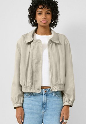 Frau trägt eine hellbeige Reißverschlussjacke mit Kragen, weißes Hemd, hellblaue High-Waist-Jeans und Creolen.