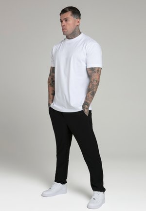 Camiseta de cuello redondo blanca, ajuste slim, combinada con pantalones de chándal ajustados negros, finalizada con zapatillas blancas. Tatuajes visibles en la piel de los brazos.
