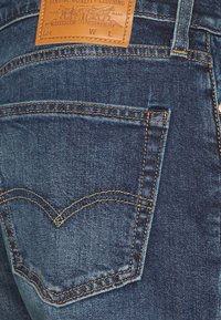 Jeans in denim blu scuro con una distintiva tasca posteriore e cuciture gialle. Presentano una patch in pelle color cuoio con il marchio sulla vita.