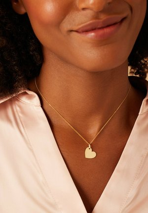 Gouden hart hanger ketting op een dunne ketting, gedragen over een lichtroze zijden blouse, die de gladde textuur en het eenvoudige ontwerp benadrukt.