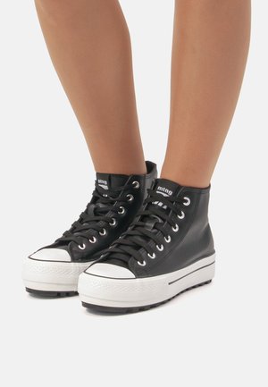 Schwarze High-Top-Sneaker mit weißen Plateausohlen und Schnürsenkeln, getragen an nackten Beinen vor einem schlichten Hintergrund.