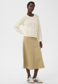 Moteris vilki kreminės spalvos megztinį su pynėmis, užsegamą sagomis, smėlio spalvos midi sijoną ir baltus sportbačius su juodomis juostelėmis, stovinti su viena ranka ant klubų.