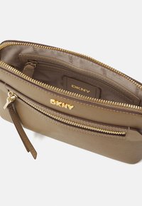 DKNY šviesiai ruda kryžminė rankinė su dviem užtrauktukais, auksinio tono detalės ir lygi tekstūra. Viduje yra audinių pamušalas ir logotipas.