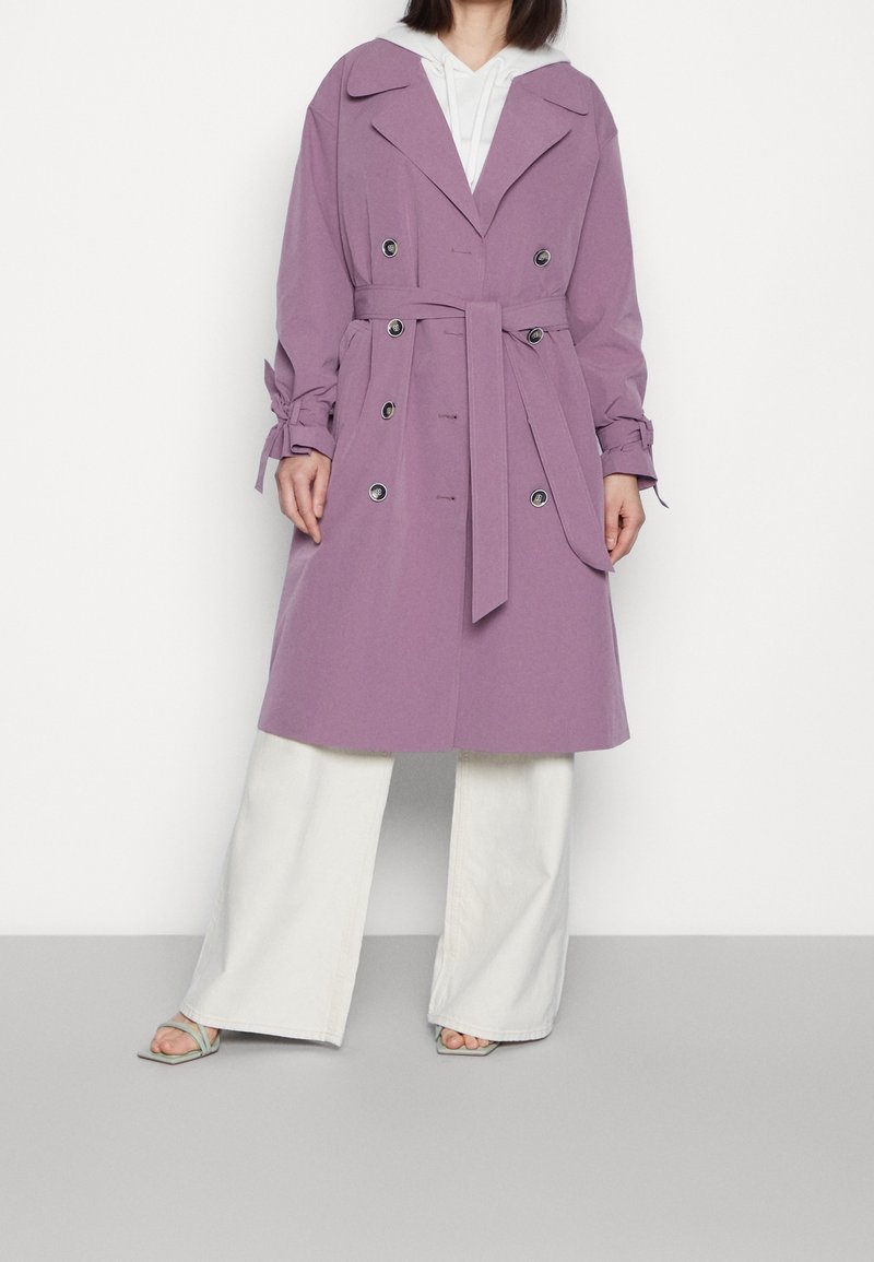 Lavendel trenchcoat med belagd midja, dubbelknäppt knäppning, breda slag och veckade ärmar, tillsammans med krämfärgade byxor.