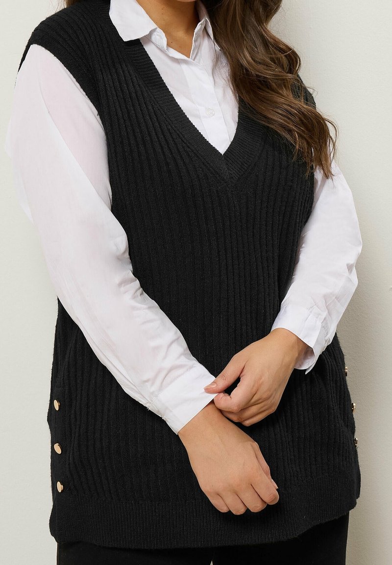 Femme portant une chemise blanche boutonnée sous un pull sans manches noir côtelé avec des boutons dorés sur le côté, les mains jointes.