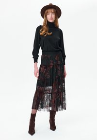 Vrouw met een bruine hoed, zwarte coltrui, doorschijnende zwarte rok met bruine bloemmotieven en bruine suède enkellaarzen, staand.