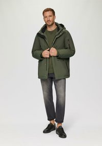 Grüne gepolsterte Jacke mit Kapuze, schwarzem Innenfutter und Seitentaschen, kombiniert mit einem dunkelgrünen Pullover, grauen Jeans und schwarzen Sneakers.