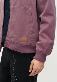 Άνδρας που φοράει ξεθωριασμένο μωβ ζακέτα με φερμουάρ και μεγάλη μπροστινή τσέπη, καθώς και μαύρο τζιν παντελόνι διακοσμημένο με ασημένια καρφιά στο μηρό.