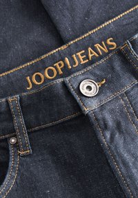 Jeans de ganga azul escuro com o logótipo dourado "JOOP!JEANS", costura visível e um botão prateado. Apresenta um design clássico de cinco bolsos.