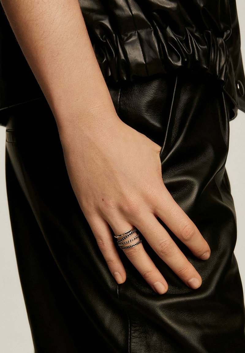 Ropa de cuero negra con una mano que lleva anillos metálicos apilados; los anillos presentan patrones lisos y texturizados, resaltando los contrastes del diseño.