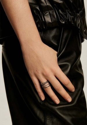 Ropa de cuero negra con una mano que lleva anillos metálicos apilados; los anillos presentan patrones lisos y texturizados, resaltando los contrastes del diseño.