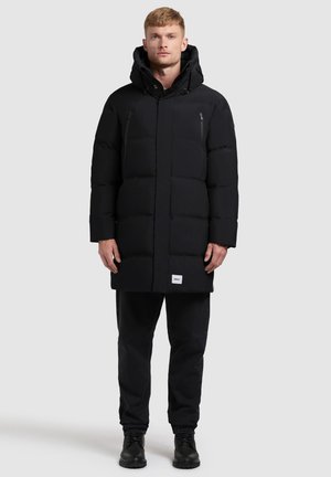 Hombre vestido con un abrigo largo acolchado negro con capucha, pantalones negros y botas negras, de pie contra un fondo gris claro.