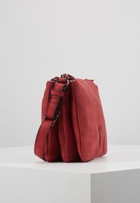 Sac à épaule en cuir perforé rouge avec sangle réglable, trois compartiments principaux et logo embossé subtil sur une surface blanche lisse.