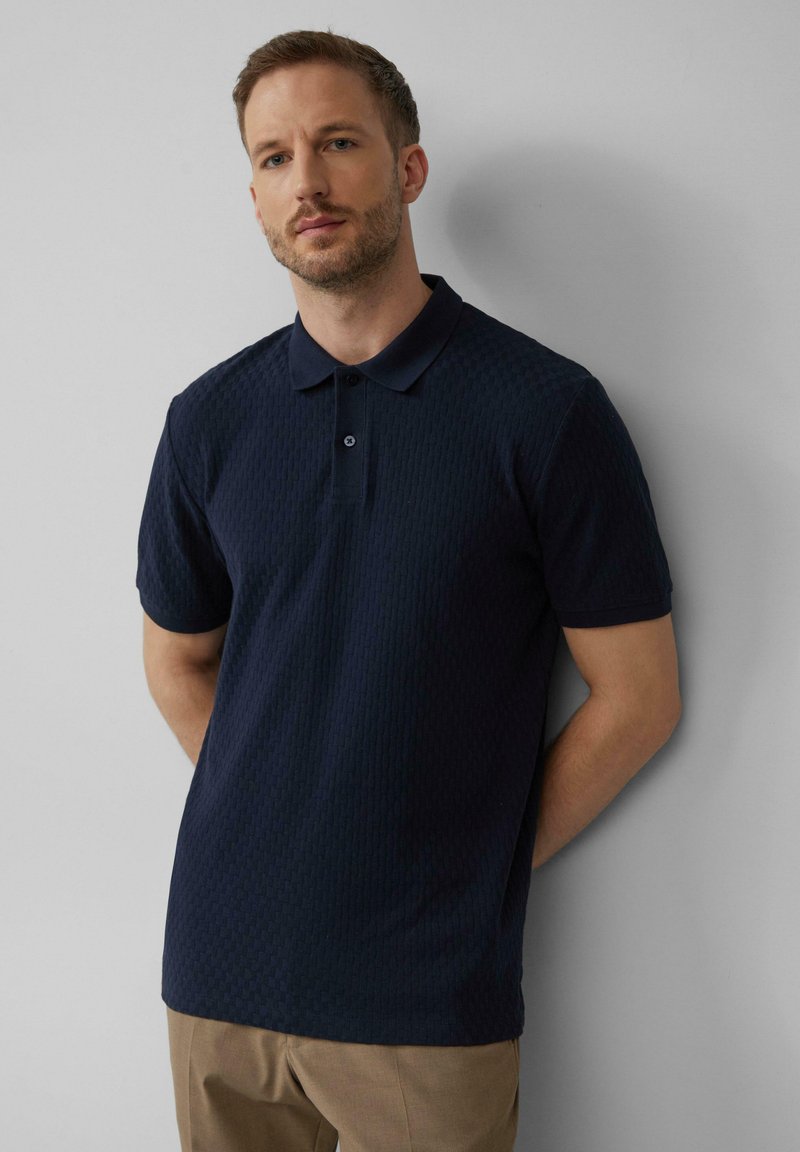 Marineblauw poloshirt met een textuurpatroon, korte mouwen en een zachte kraag. Voorzien van twee knopen bij de hals en een losse pasvorm.