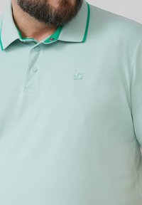 Lichte mintgroene polo met een gestructureerde stof, contrasterende groene kraagafwerking, knoopsluiting met drie knopen en een verzonken logo op de borst.