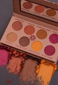 Paleta cieni do powiek z ośmioma okrągłymi pojemnikami w odcieniach pomarańczowym, brązowym, różowym i złotym, z teksturowanymi proszkami rozsypanymi poniżej.