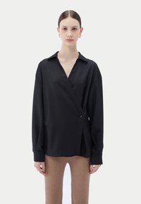 Svart omlottblus med en urgröpt krage och en knappdetalj, gjord av slätt tyg. Blusen är kombinerad med ribbstickade beige byxor.