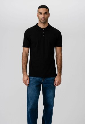 Mies seisoo kasvot kohti edessä, pukeutuneena mustaan lyhythihaiseen poolopaitaan ja sinisiin farkkuihin, yksinkertaisen vaalean taustan edessä.