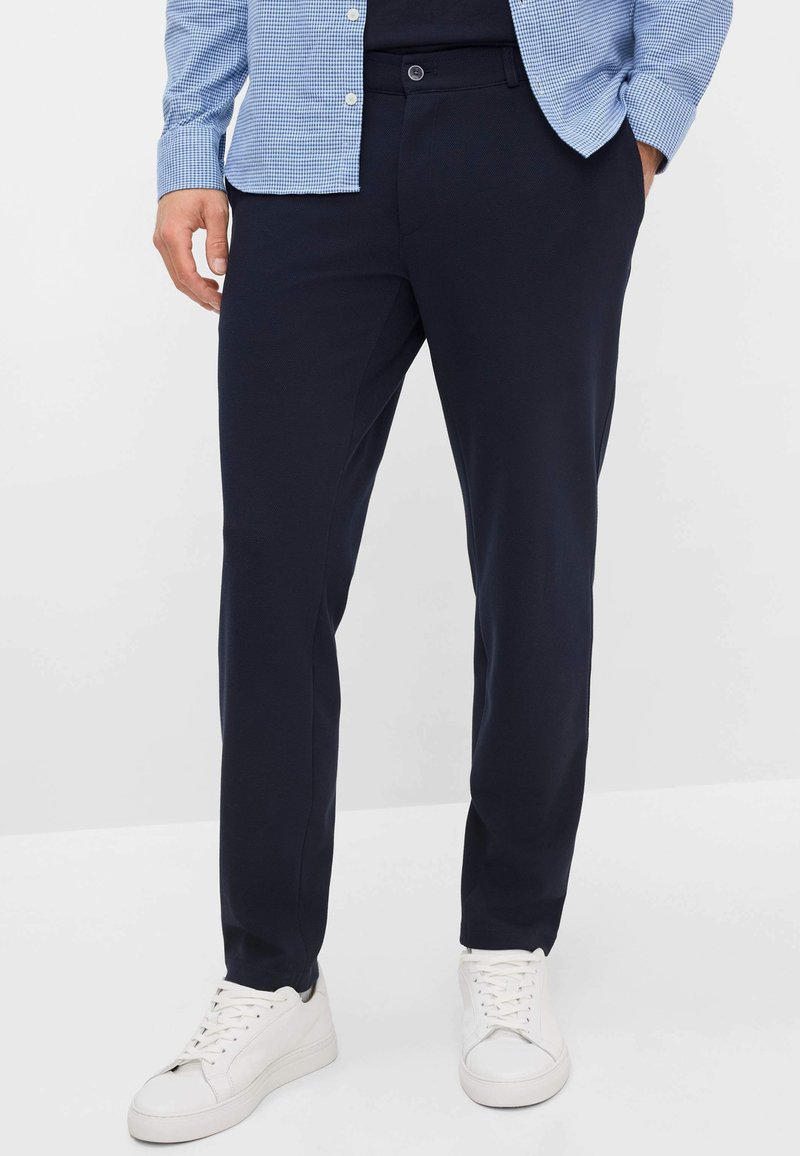 Marineblauwe broek van gladde stof, met een slanke pasvorm en een enkele knoopsluiting. Gecombineerd met witte sneakers voor contrast.