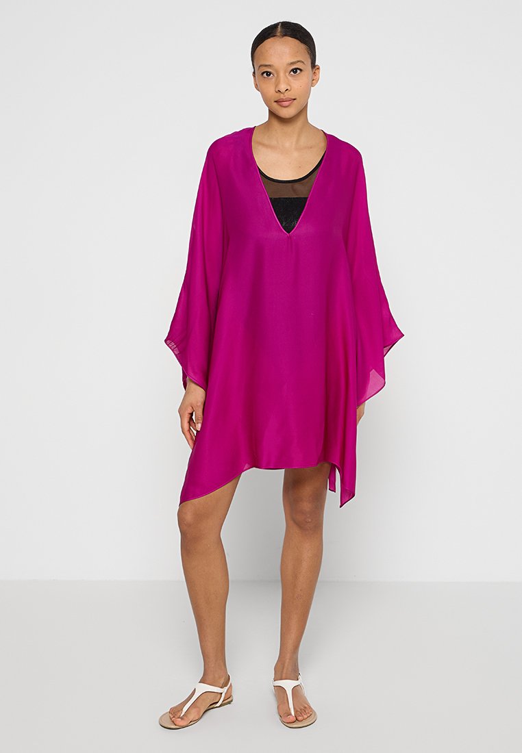 Fuchsia Kaftan mit V-Ausschnitt, weit ausgestellten Ärmeln und asymmetrischem Saum. Aus leichtem Stoff gefertigt, über einem schwarzen Top getragen. Weiße Sandalen dazu.