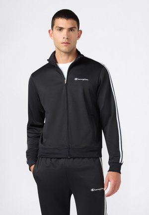 Mann trägt einen schwarzen Champion Trainingsanzug mit weißen Streifen an Ärmeln und Hosen, steht mit einer Hand in der Tasche vor einfarbigem Hintergrund.