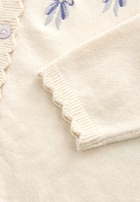 Cream cardigan made of soft fabric, featuring floral embroidery in blue tones and a scalloped edge on the cuff. Rounded buttons displayed.