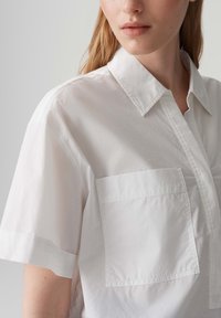 Weiße Bluse mit kurzen Ärmeln und kragenförmigem Ausschnitt, ausgestattet mit einer einzelnen Brusttasche. Der Stoff hat eine glatte Textur und einen dezenten Glanz.