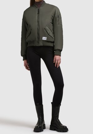 Person wearing a green bomber jacket, black leggings, and black chunky boots standing against a plain background.